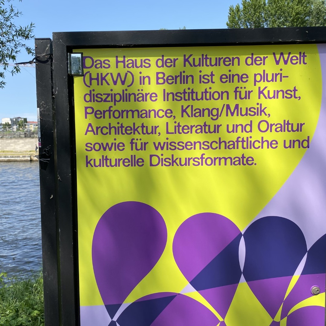Plakat an der Spree in Berlin mit der Aufschrift: Das Haus der Kulturen der Welt (HKW) ist eine pluridisziplinäre Institution für Kunst, Performance, Klang/Musik, Architektur, Literatur und Oraltur sowie für wissenschaftliche und kulturelle Diskursformate.